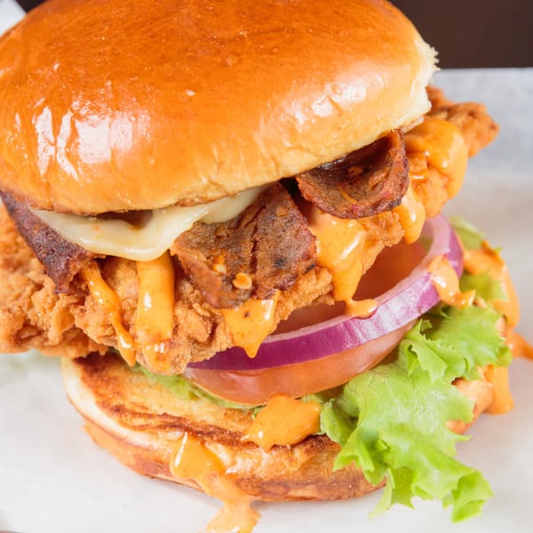 A crispy chicken sandwich with bacon, melted cheese, red onion, lettuce, and orange sauce.